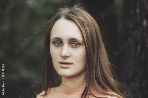 Portrait of a young woman with neutral face expression looking to the camera, with a forest background