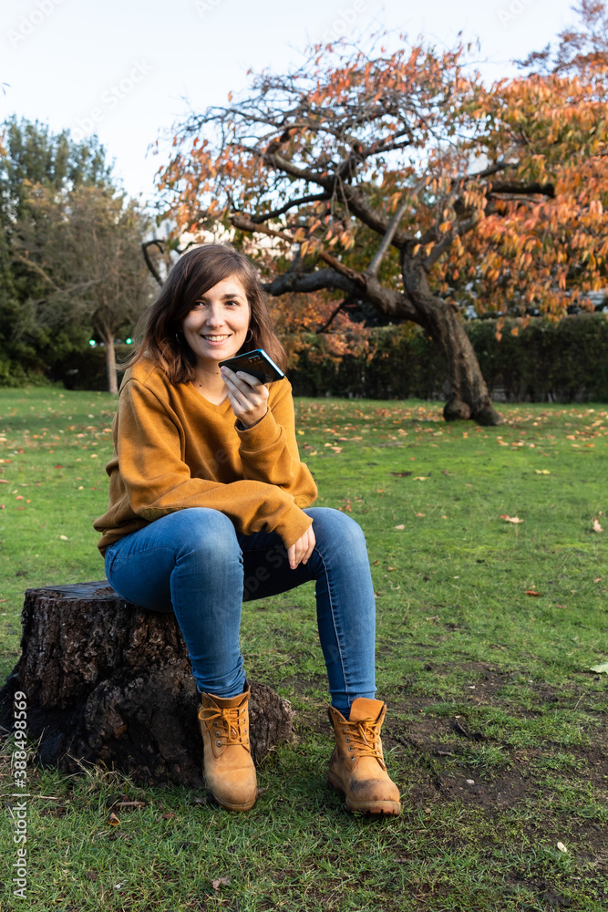 Attractive young female sitting on the base of an ancient tree that has been cut and sending a recording to a friend with her smartphone at Regents Park during the autumn season.