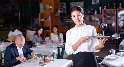 Hospitable attractive waitress with serving tray meeting guests in cozy restaurant..