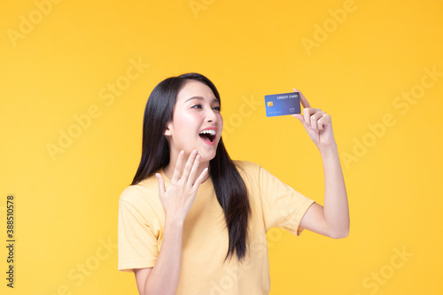 Cheerful young asian woman in yellow T-shirt holding bank card isolated on yellow background