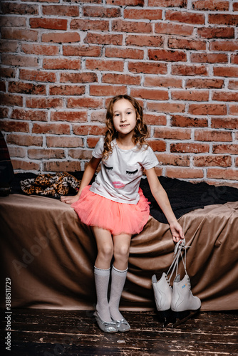 A little girl in a pink skirt sits on a bed against a background of a brick wall, next to white skates. Children's style and fashion concept. Long white knee-highs.