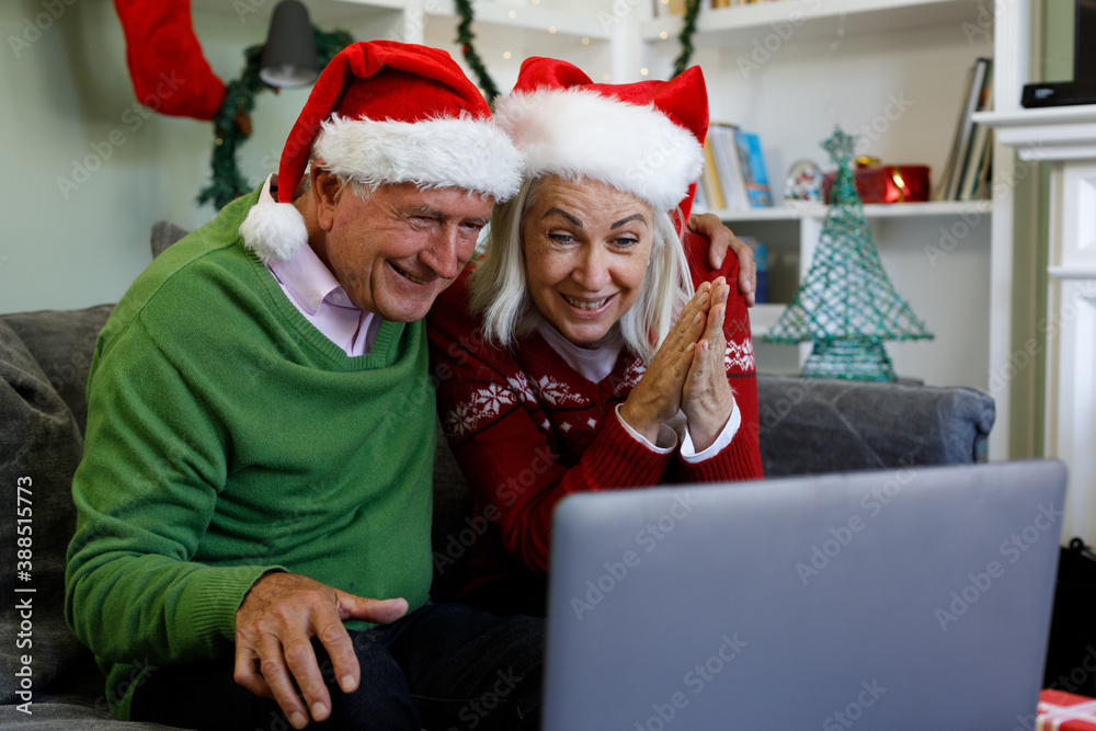 Senior couple in santa hat sitting on couch smiling while having a video call on laptop at home Senior couple in santa hat sitting on couch smiling while having a video call on laptop at home