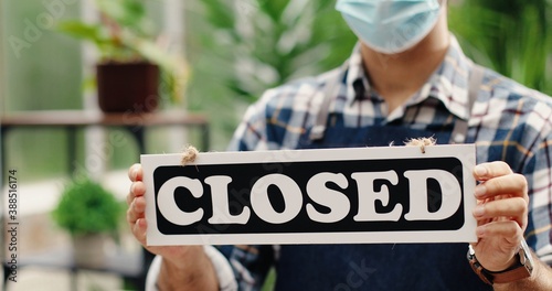 Close up of Caucasian man hands holding Closed sign indoors in florist shop in quarantine. Young male flower center employee in apron and mask standing in garden center with sign. Business concept
