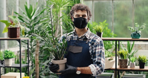 Close up portrait of happy handsome Caucasian man employee in apron and mask holding pot with herb and smiling to camera. Joyful young male flower shop worker with plants in good mood Business concept