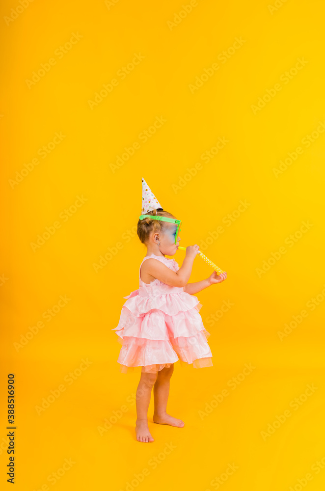 a little girl stands sideways with big glasses and a holiday hat blowing a whistle on a yellow background with space for text a little girl stands sideways with big glasses and a holiday hat blowing a whistle on a yellow background with space for text