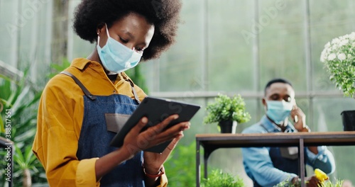 Portrait of beautiful African American female worker in mask tapping on tablet in flower shop in foreground. Handsome young male employee florist speaking on smartphone on background. Job concept