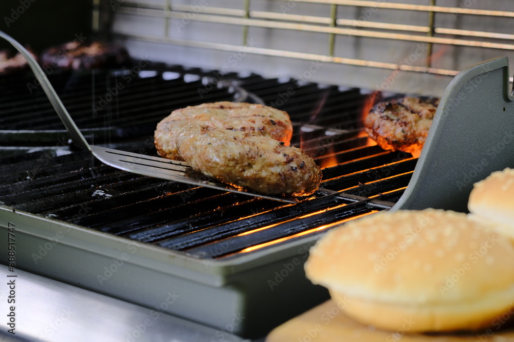 The process of cooking beef patties for burgers on a gas grill. Turkey and Beef cutlets cooked on a gas grill outside. In the foreground Burger buns with sesame seeds