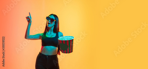 Watching cinema with popcorn, pointing up. Young caucasian woman's portrait on gradient yellow-orange studio background in neon. Concept of youth, human emotions, facial expression, sales, ad. Flyer