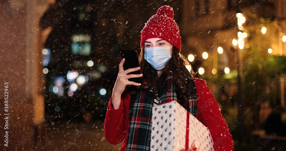 Portrait of happy beautiful young woman typing on smartphone while standing outside on snowy street. Joyful female with present texting on mobile phone outdoors in good mood. Winter concept