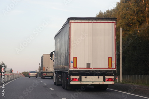 European heavy trucks with white semi trailers van drive on two lane suburban asphalted highway road, back view at summer evening on forest and sky background, transportation logistics
