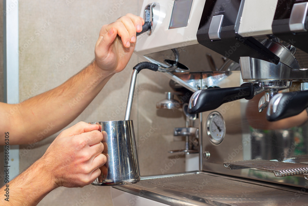 Professional barista holding metal jug warming milk using the coffee machine. Young man preparing coffee at counter. Professional barista holding metal jug warming milk using the coffee machine. Young man preparing coffee at counter.