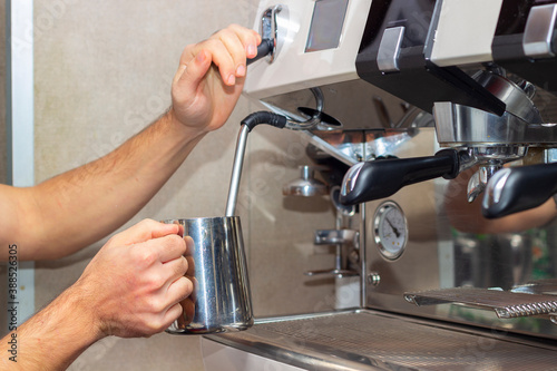 Professional barista holding metal jug warming milk using the coffee machine. Young man preparing coffee at counter. © mestock