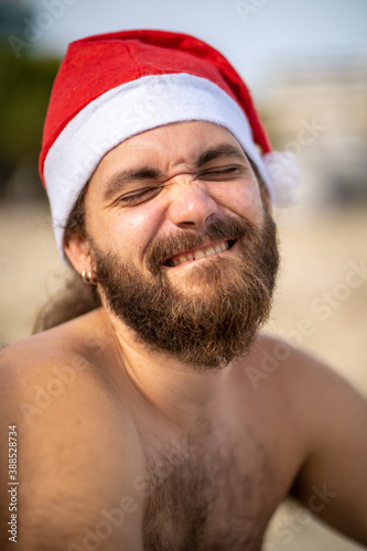 Vertical portrait of a bearded man with a Santa Claus hat expressing joy to camera, with his eyes closed in a beach.