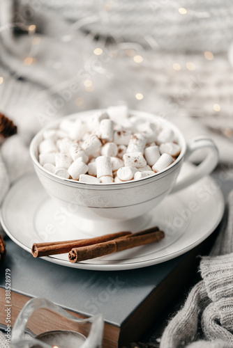 Still life details, cup of coffee in living room, top view point. Winter weekend with a book on the sofa. White knitted sweaters are stacked. Delicate marshmallow with cinnamon in a mug with coffee