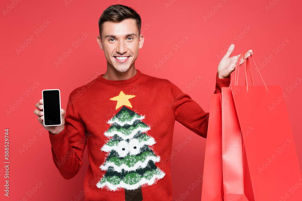 Smiling man in sweater with pine tree holding smartphone with blank screen and shopping bags on red background