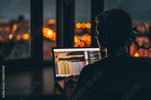 A young male programmer in front of a window with a lights of night city, on his laptop screen is code. out of focus photo