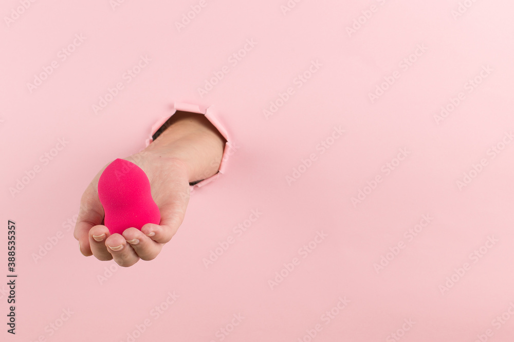 Girl hand holds a beauty blender for makeup from a hole in a pink background, copy space.