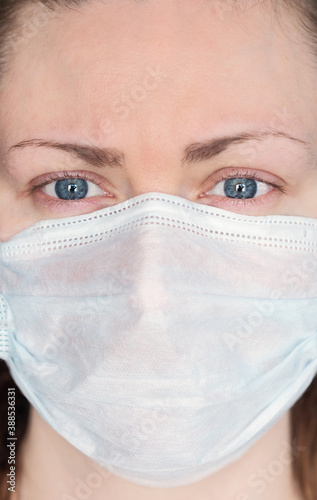 Close up face portrait of young woman with blue eyes wearing a surgical mask to protect herself and avoid the spread of covid-19 coronavirus and looking straight at camera - viewer photo