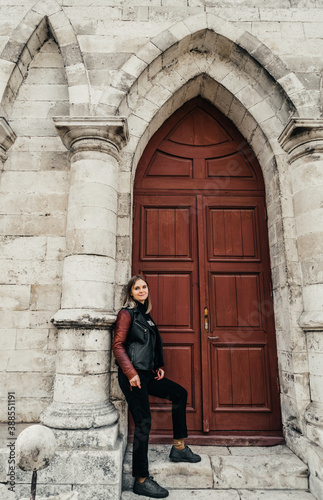 young woman in a leather jacket near the entrance to the ancient wooden door of a gothic temple