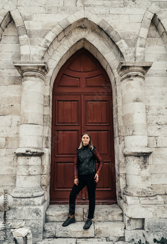 young woman in a leather jacket near the entrance to the ancient wooden door of a gothic temple