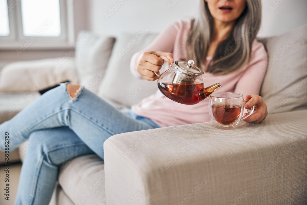 Middle-aged woman sitting on couch and pouring tea into cup Middle-aged woman sitting on couch and pouring tea into cup