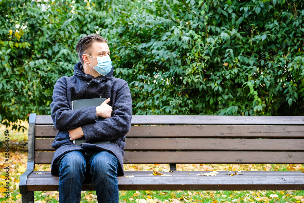 Fototapeta premium Handsome young European man in a park with laptop with a medical face mask on. Freelance working outside the office during an Covid-19 epidemic. Selective focus