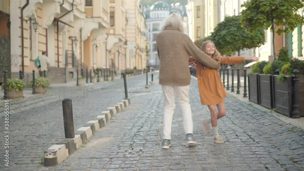 Wide shot portrait of cheerful senior man and cute little girl jumping and dancing on city street. Happy carefree grandfather and granddaughter having fun on sunny day outdoors and leaving. Wide shot portrait of cheerful senior man and cute little girl jumping and dancing on city street. Happy carefree grandfather and granddaughter having fun on sunny day outdoors and leaving.
