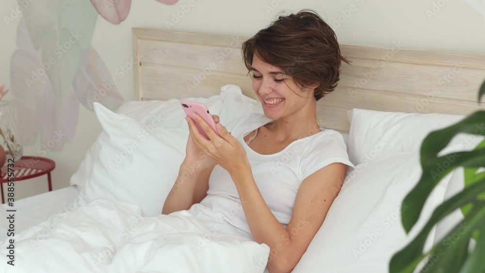 Dreamful calm young woman in white t-shirt pajamas home wear lying in bed spending time in bedroom hold in hand using mobile cell phone send typing sms message. Rest relax good mood lifestyle concept Dreamful calm young woman in white t-shirt pajamas home wear lying in bed spending time in bedroom hold in hand using mobile cell phone send typing sms message. Rest relax good mood lifestyle concept