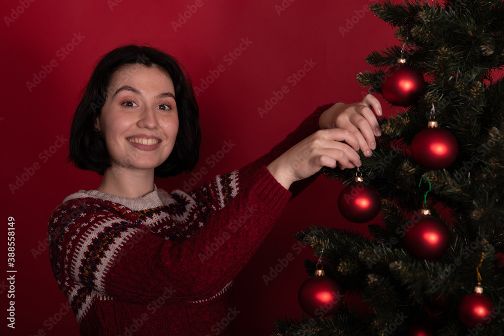 Happy pretty woman in sweater with ornament decorating xmas tree looks at camera with smile on red background. Christmas New Year's Eve preparation, holiday stress-free planning, festive decor concept Happy pretty woman in sweater with ornament decorating xmas tree looks at camera with smile on red background. Christmas New Year's Eve preparation, holiday stress-free planning, festive decor concept