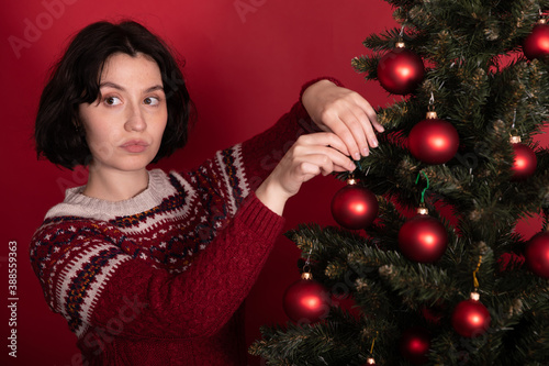 Obraz na płótnie Serious pretty woman in sweater with ornament decorating xmas tree looks to side, red background
