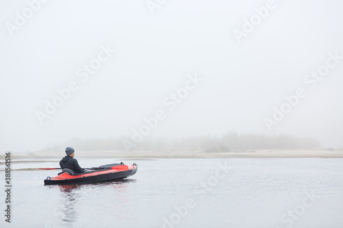 Rear view of handsome man sitting in canoe and looking in distance, guy wearing black jacket and cap, foggy autumn morning, sportsman padding in lake.