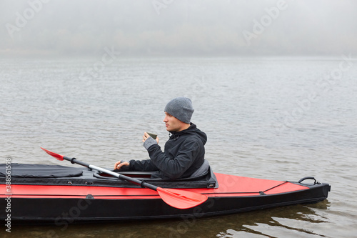 Man paddles red and black kayak and sitting with paddle, holds mug with hot coffee or tea on river or lake in fall season. Autumn kayaking, water extreme sport.