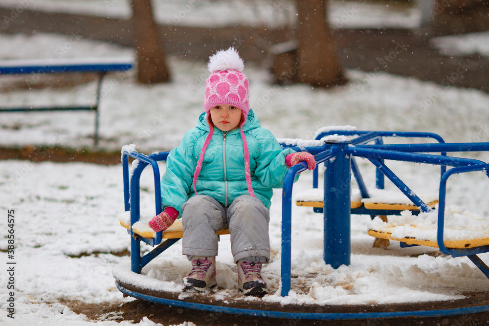 Little sad girl sitting on a carousel on a snowy winter day. The girl is not happy about the first snow
