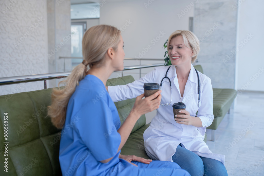 Two smiling female medical workers sitting on sofa, drinking coffee Two smiling female medical workers sitting on sofa, drinking coffee