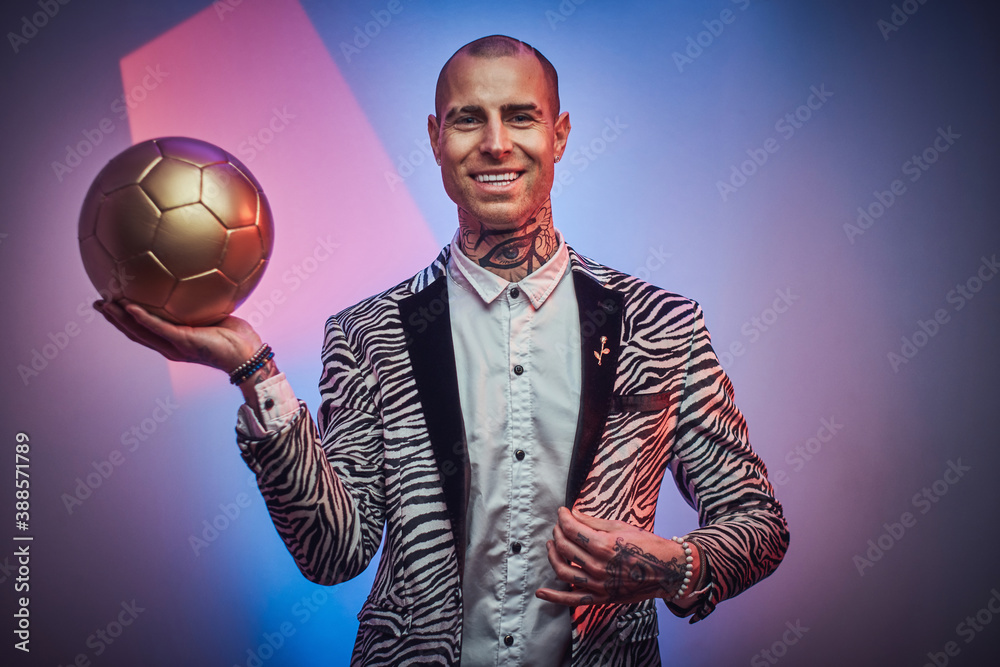 Plakat Joyful tattooed guy with jewellery in stylish suit poses in colored abstract background holding golden ball.