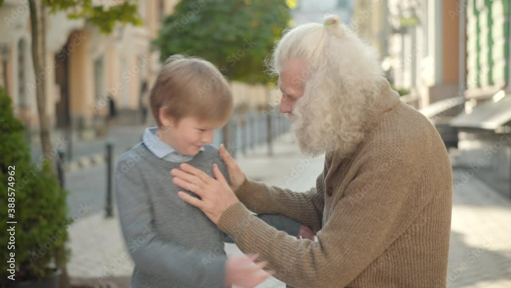 Caring old Caucasian man adjusting shirt of smiling little boy on city street. Thankful grandson hugging grandfather and leaving. Happy grandparent and grandchild on sunny day outdoors. Caring old Caucasian man adjusting shirt of smiling little boy on city street. Thankful grandson hugging grandfather and leaving. Happy grandparent and grandchild on sunny day outdoors.