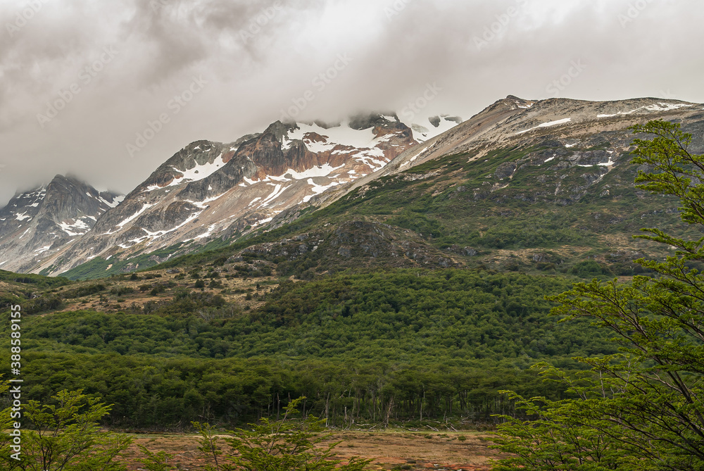Ushuaia, Tierra del Fuego, Argentina - December 13, 2008: Martial Mountains in Nature Reserve. Brown snow covered mountain peaks rising into brown cloudscape. Green forest in front.