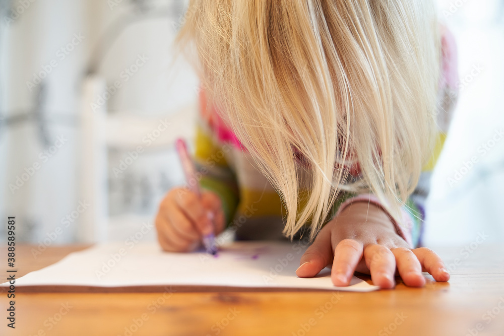 4 year old blonde girl with a brightly colored striped sweater sits at a wooden table and draws with a purple pencil on a piece of white paper