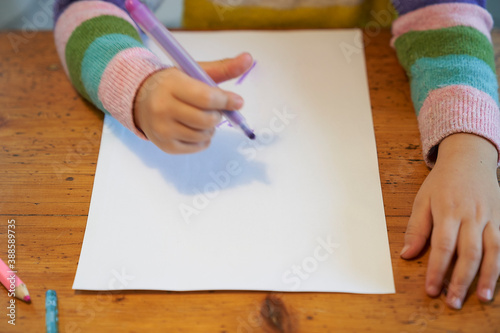 4 year old blonde girl with a brightly colored striped sweater sits at a wooden table and draws with a purple pencil on a piece of white paper