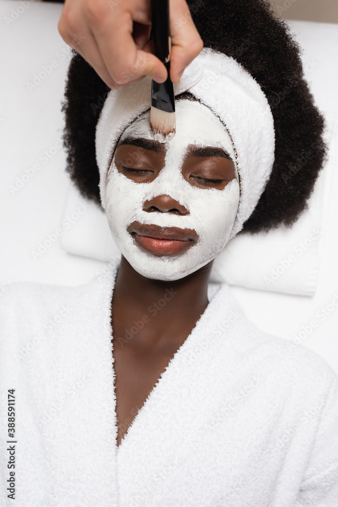 top view of positive african american woman wearing bathrobe, lying near spa therapist applying face mask on forehead in spa salon top view of positive african american woman wearing bathrobe, lying near spa therapist applying face mask on forehead in spa salon