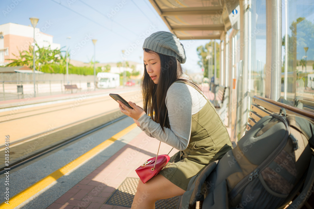 outdoors lifestyle portrait of young beautiful and happy Asian Korean woman waiting the train sitting on station platform bench using mobile phone checking schedule online
