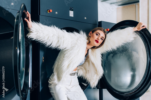 young trendy woman in faux fur jacket standing near washing machines in public laundromat photo