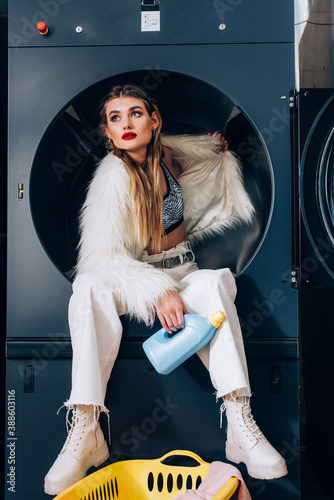 young woman in white faux fur jacket sitting in washing machine and holding bottle with detergent in laundromat