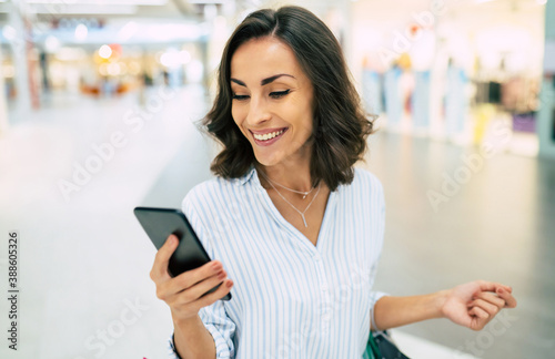 Modern gorgeous young smiling woman in trendy stylish clothes with bright colorful shopping bags is using her smart phone while walking in the mall