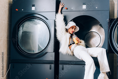 trendy woman in faux fur jacket and hat holding plastic cup and drinking orange juice near washing machines in public laundromat photo