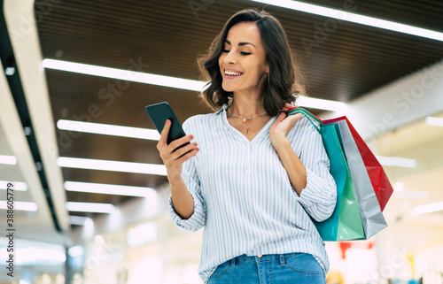 Modern gorgeous young smiling woman in trendy stylish clothes with bright colorful shopping bags is using her smart phone while walking in the mall photo