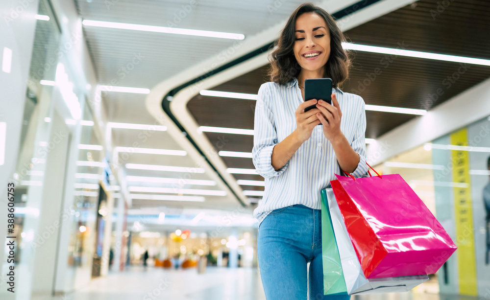 Modern gorgeous young smiling woman in trendy stylish clothes with bright colorful shopping bags is using her smart phone while walking in the mall Modern gorgeous young smiling woman in trendy stylish clothes with bright colorful shopping bags is using her smart phone while walking in the mall