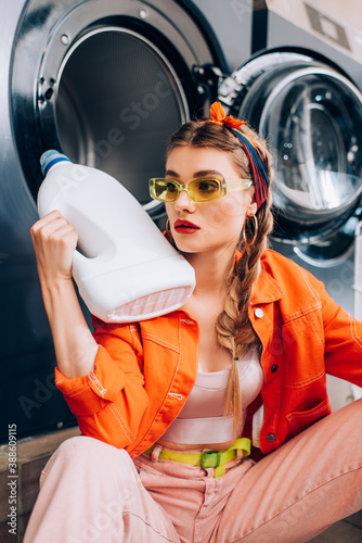 young woman in sunglasses holding bottle with detergent in laundromat photo