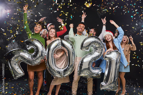 Group of young excited multiracial friends holding silver foil balloons in form of numbers 2021, raising hands up while celebrating New Years Eve, confetti falling in the air