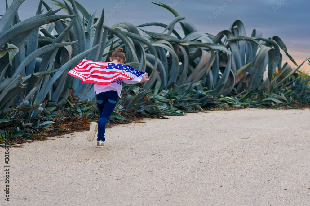 Φωτογραφία Back view little girl holding a flag of America, running on the road at sunset
